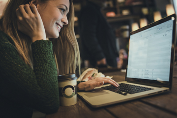 A woman smiling and looking at her laptop with a coffee in front of her.